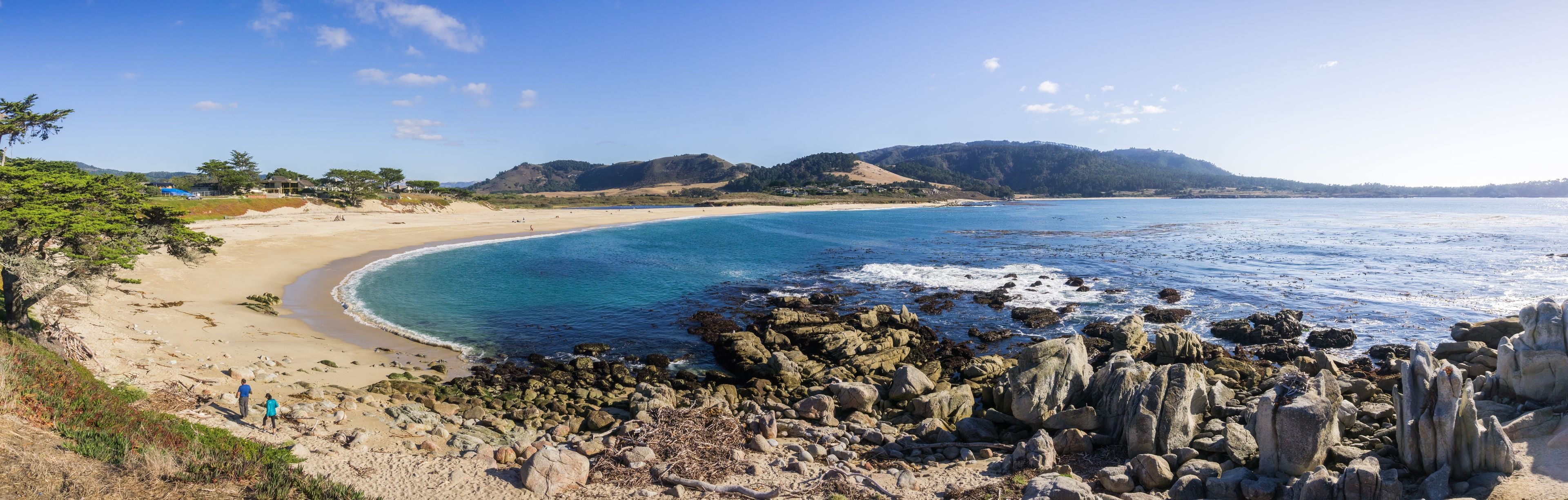 Paesaggio costiero con spiaggia sabbiosa, acque blu cristalline, rocce affioranti e colline in lontananza.