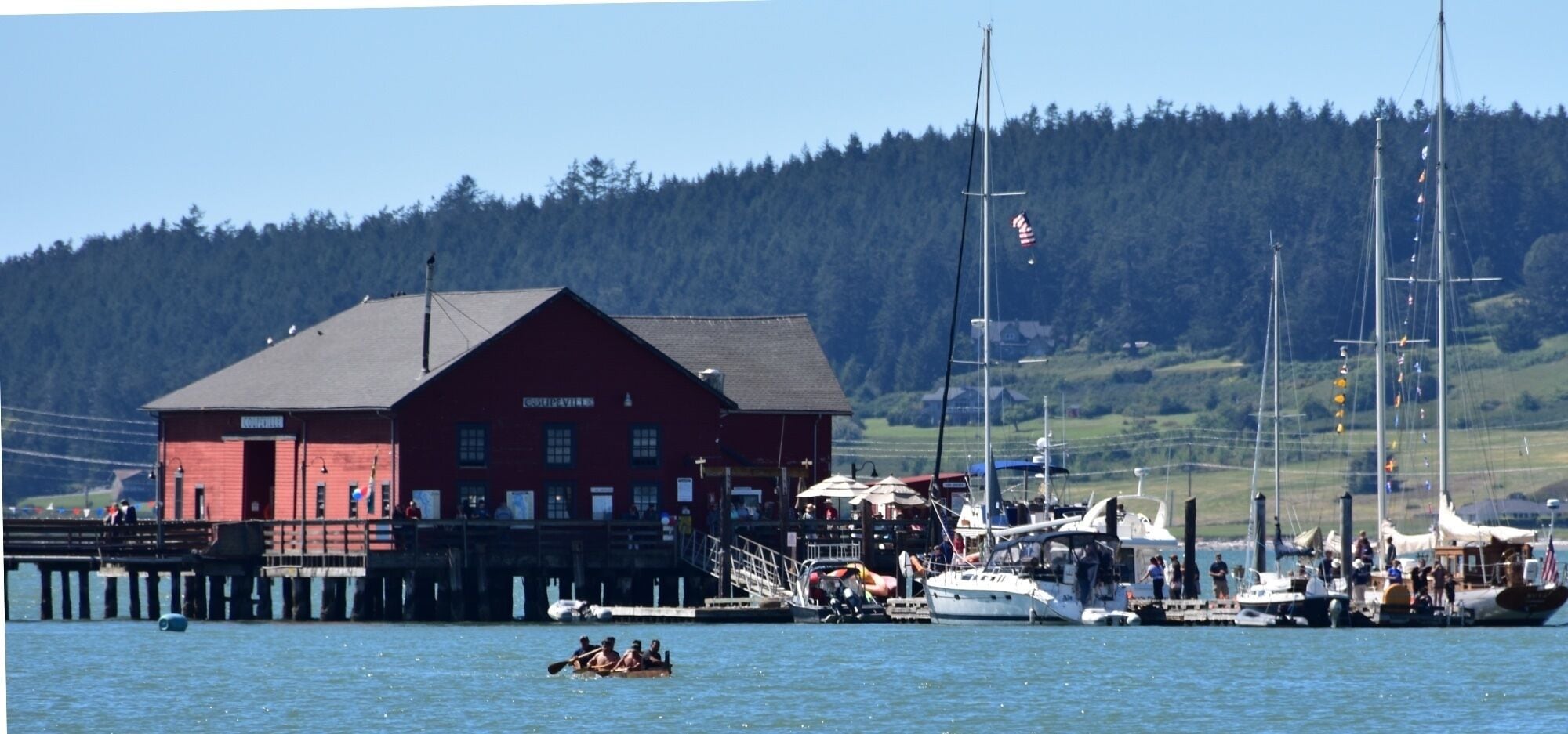 A red building with a dock extending into the water, surrounded by boats and a forested hillside.