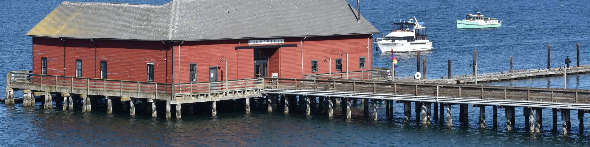 A red building on stilts by a calm body of water with a boat docked nearby.