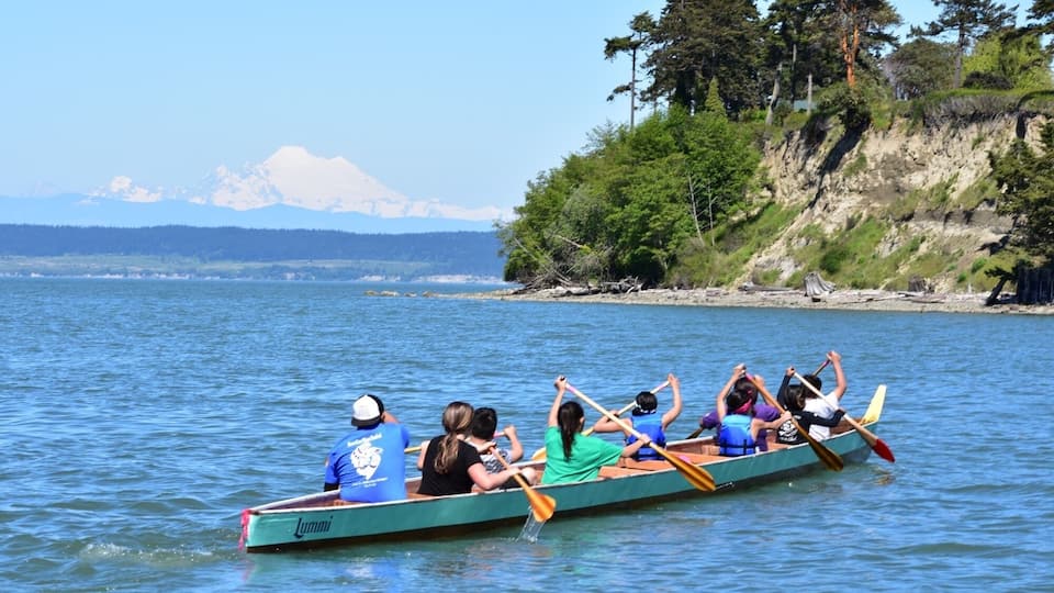 A rowing team in a green boat on a calm lake with a forested shore and mountains in the background.