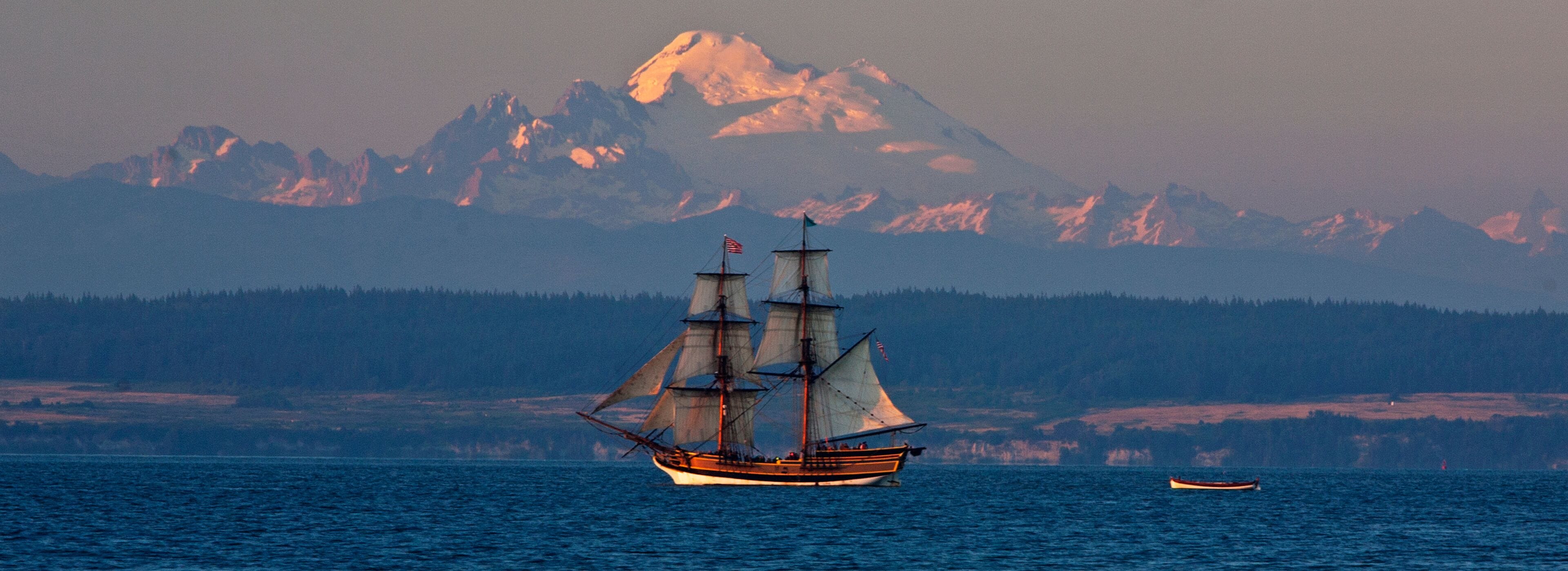 A sailing ship on calm waters with a mountain range in the background.