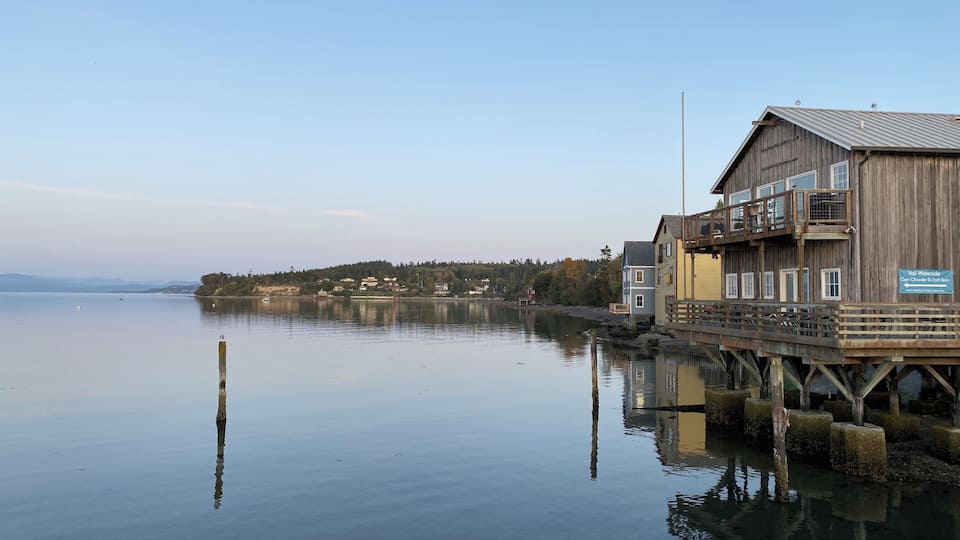A calm lake with a wooden pier extending into the water, a wooden building with a sign, and a distant shoreline with houses and trees.