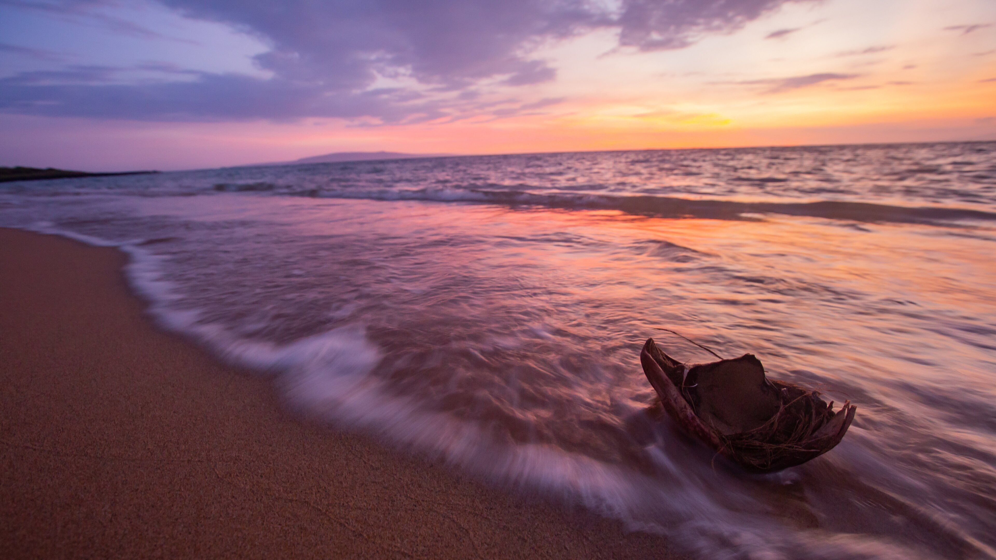 A beach at sunset with waves gently lapping the shore and a boat on the sand.
