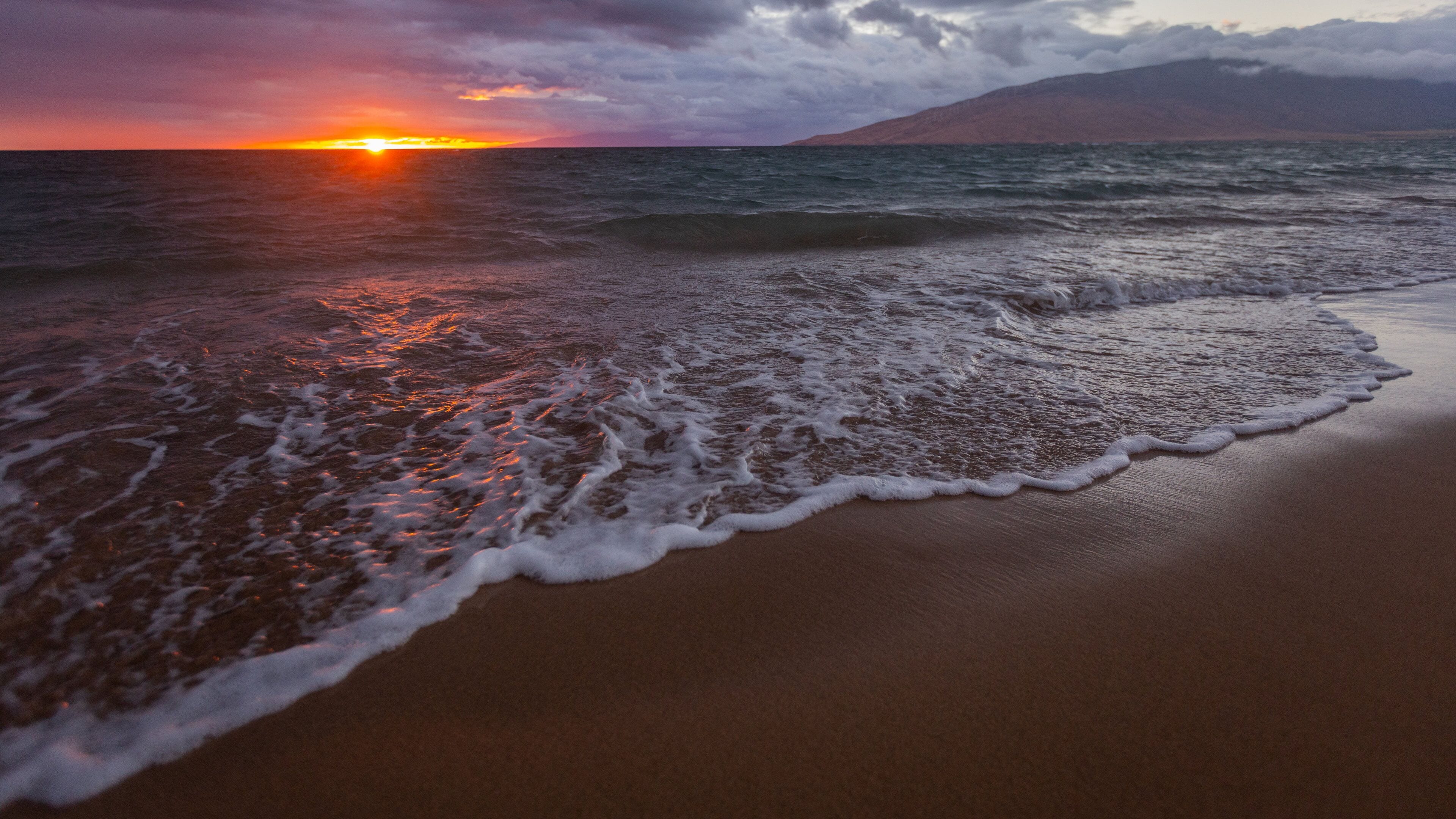 A beach with waves at sunset, a mountain in the background, and a cloudy sky.