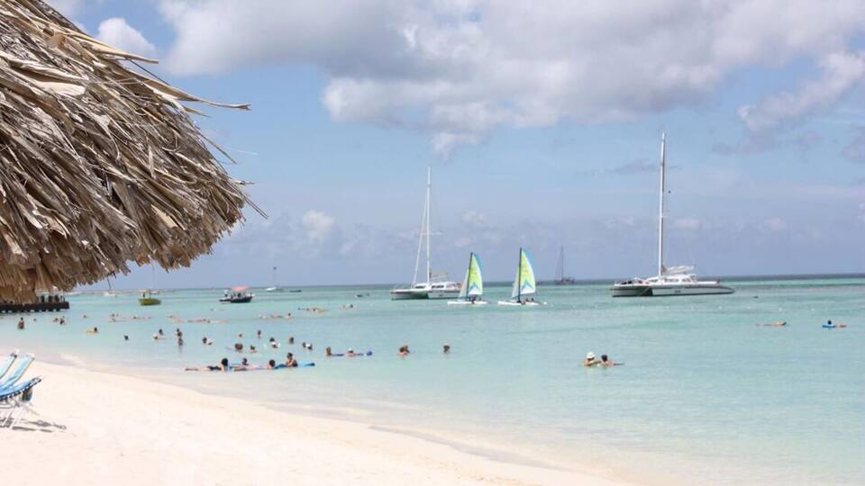A beach with clear turquoise water, sailboats, and people swimming and sunbathing.