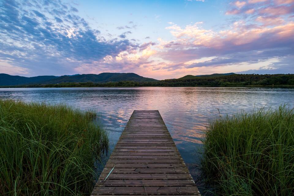 Une jetée en bois s’étendant dans un lac paisible, entourée de verdure et bordée de collines lointaines, sous un ciel partiellement nuageux.