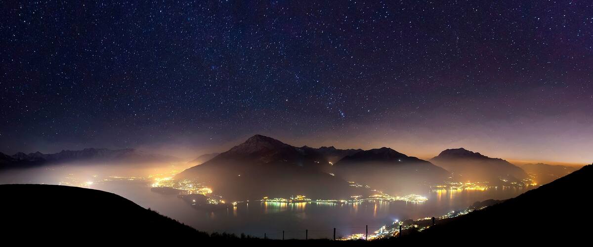 A starry night sky over a mountainous landscape with a lake and city lights reflecting on the water.