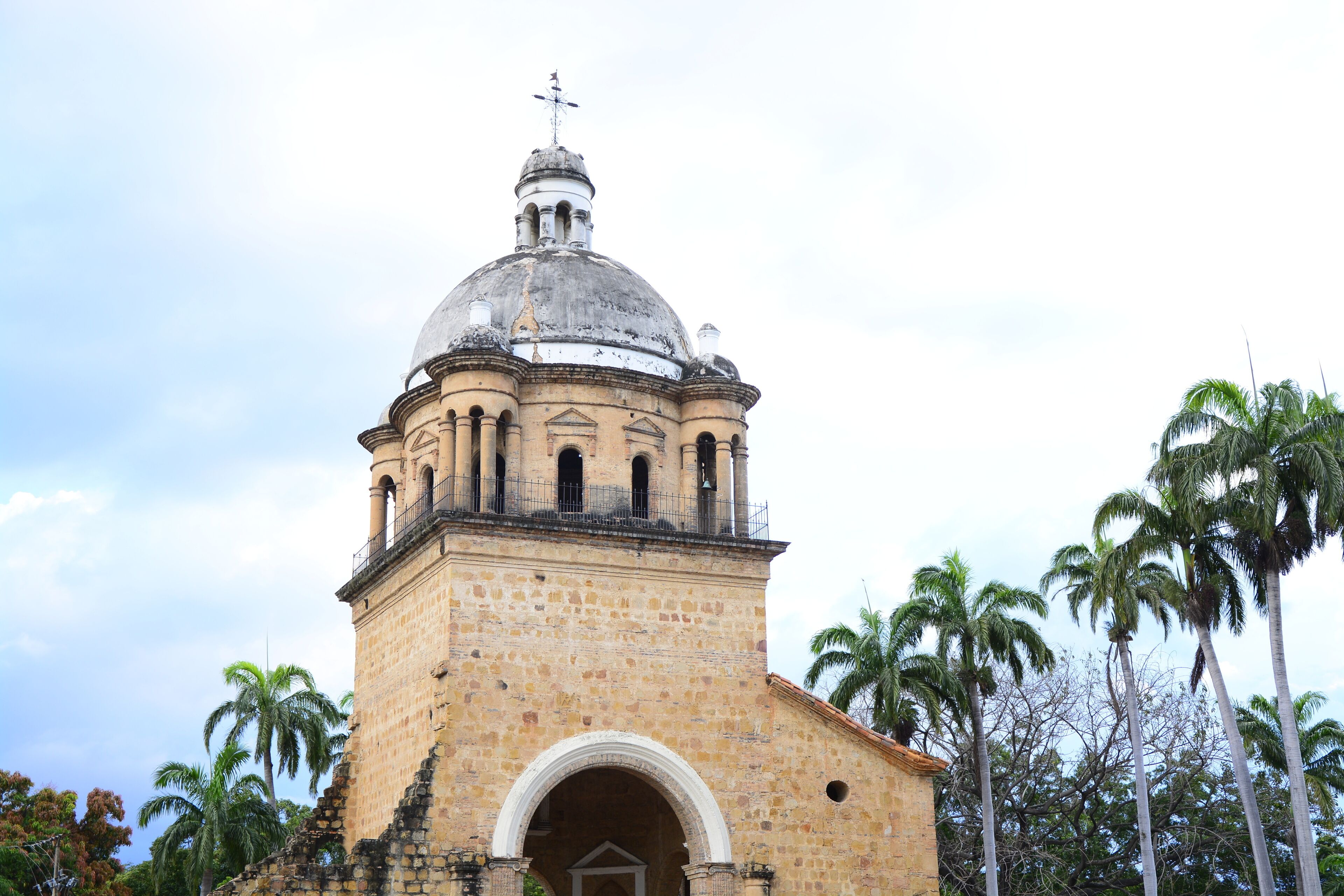 Una torre de piedra histórica con cúpula y campanario, rodeada de palmeras.