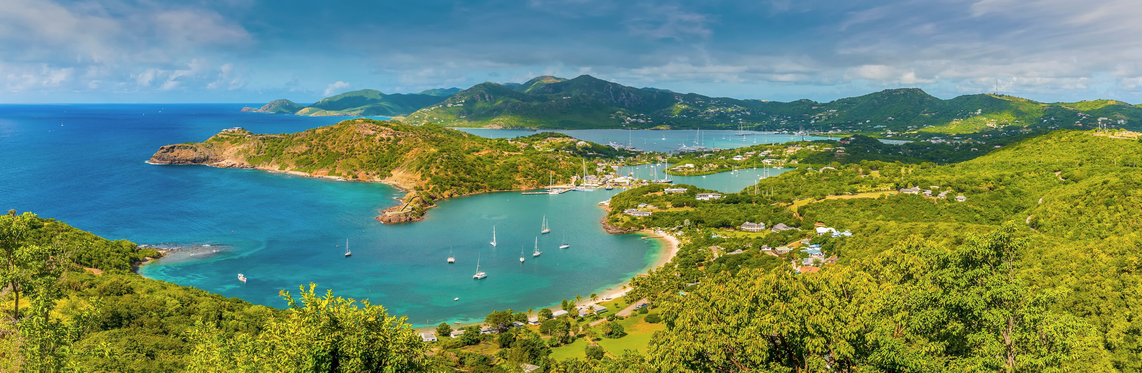 A coastal landscape with clear turquoise waters, a bay with sailboats, and lush green hills.