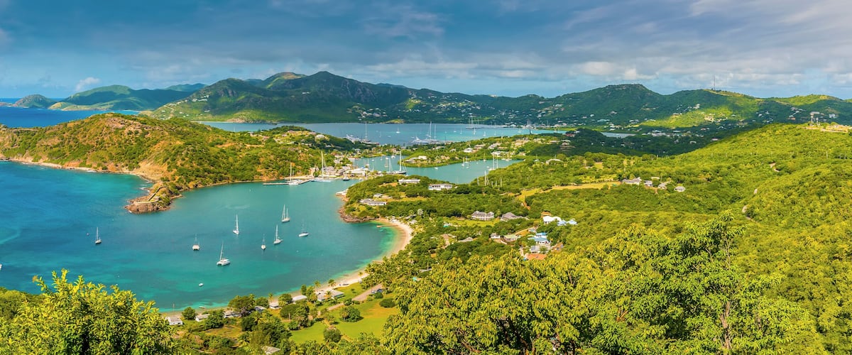 A coastal landscape with clear turquoise waters, a bay with sailboats, and lush green hills.