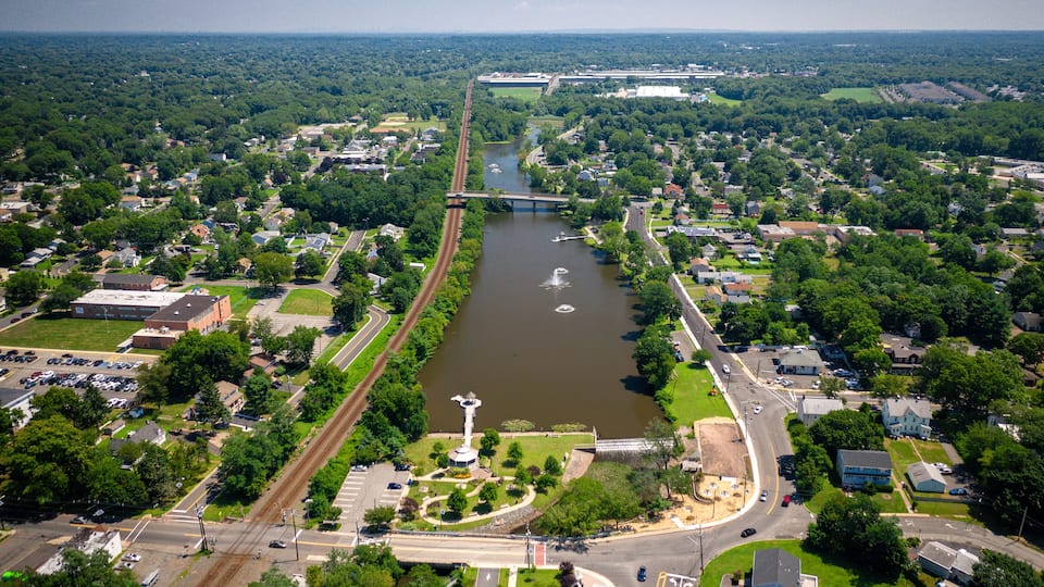 An aerial view of a river flowing through a town with a bridge, surrounded by greenery and residential areas.