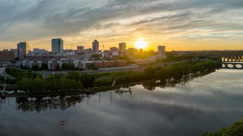 A city skyline at sunset with a calm river reflecting the buildings, a bridge with circular structures, and a clear sky.