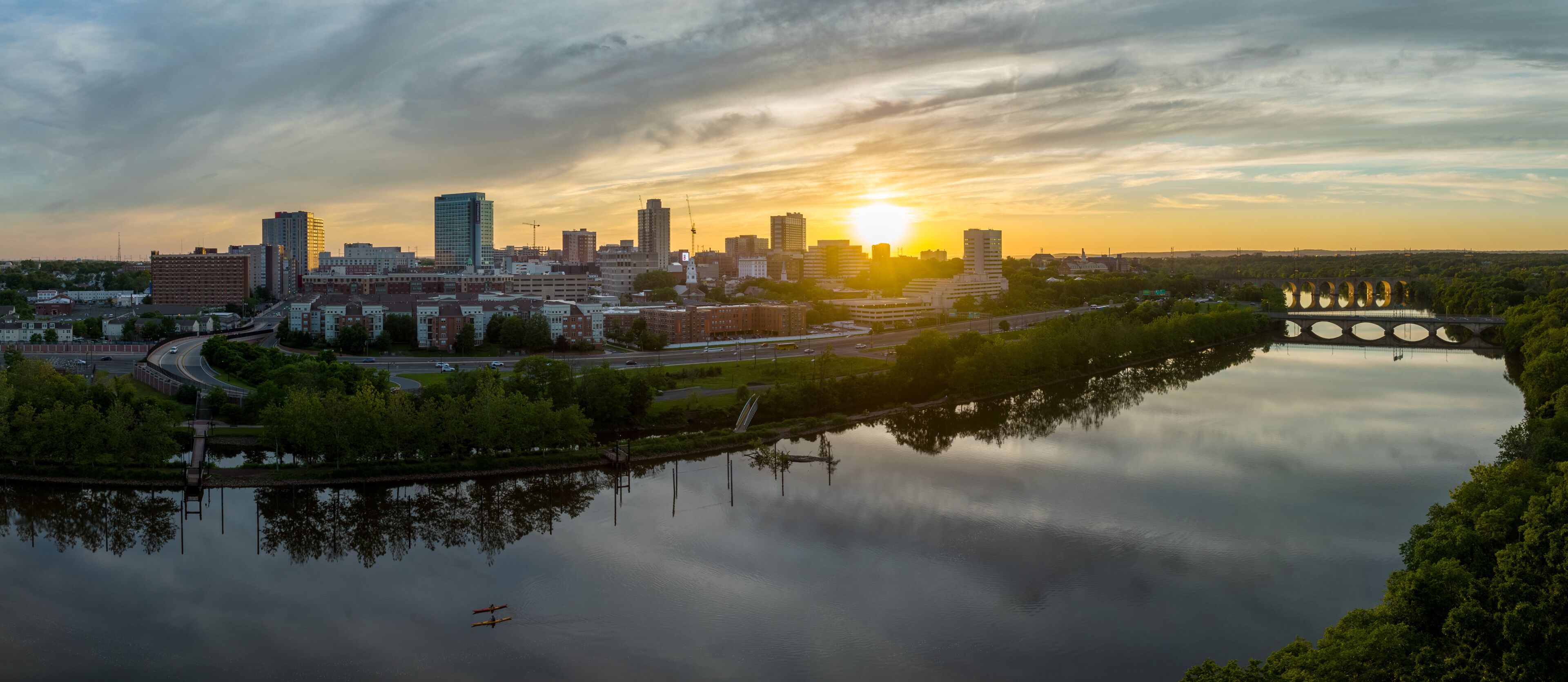 A city skyline at sunset with a calm river reflecting the buildings, a bridge with circular structures, and a clear sky.