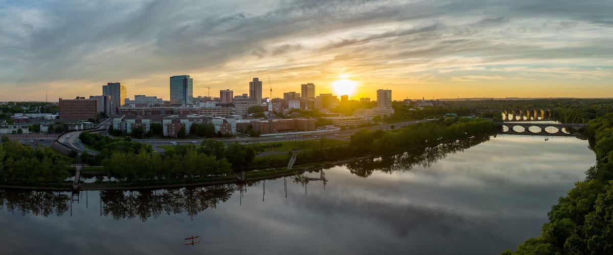 A city skyline at sunset with a calm river reflecting the buildings, a bridge with circular structures, and a clear sky.