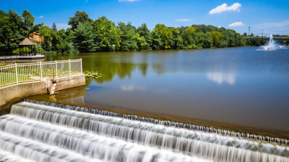 A calm lake with a waterfall, a gazebo, and lush greenery.