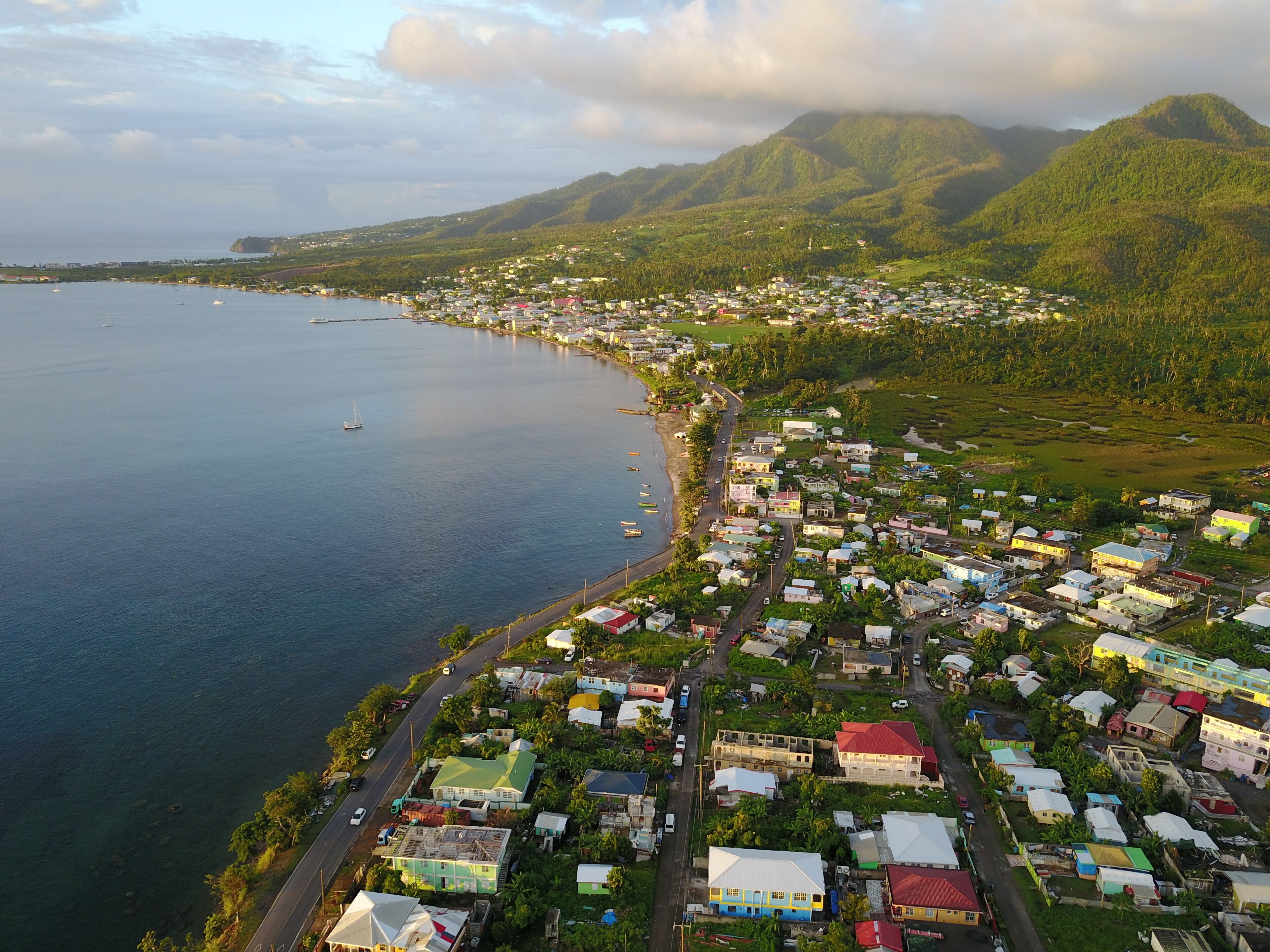 A coastal town with a dense cluster of colorful houses, a calm sea, and lush green hills in the background.