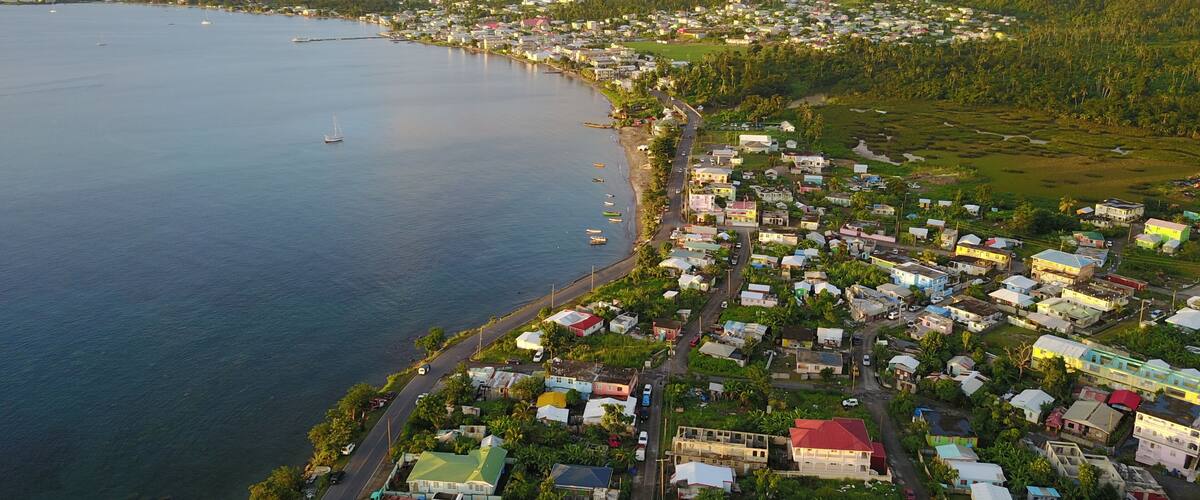 A coastal town with a dense cluster of colorful houses, a calm sea, and lush green hills in the background.