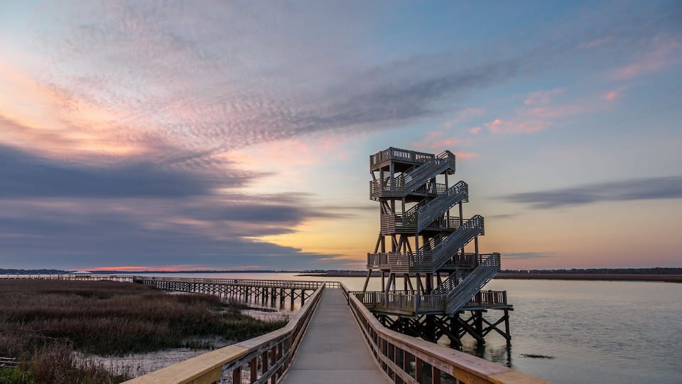 A wooden boardwalk leading to a metal staircase structure by a body of water with a sunset sky.