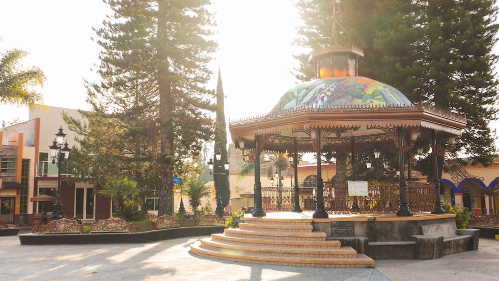 Un kiosque surmonté d’un dôme aux couleurs vives, entouré d’arbres et bordé d’une place dallée.