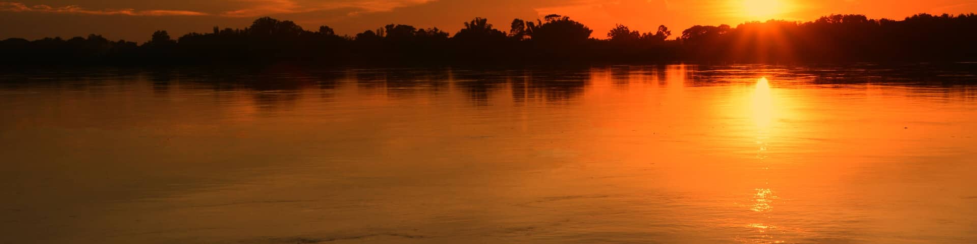 A sunset over a calm lake with a silhouette of trees and a mountain in the background.