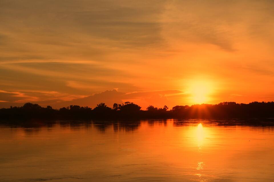 Een zonsondergang boven een rustig meer, met silhouetten van bomen en een berg op de achtergrond.
