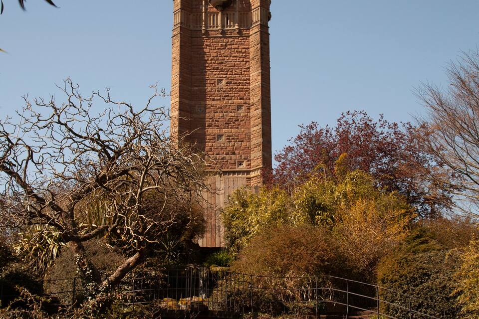 Een hoge bakstenen toren met ingewikkelde architectuur, omringd door weelderige begroeiing en een kleine waterval.