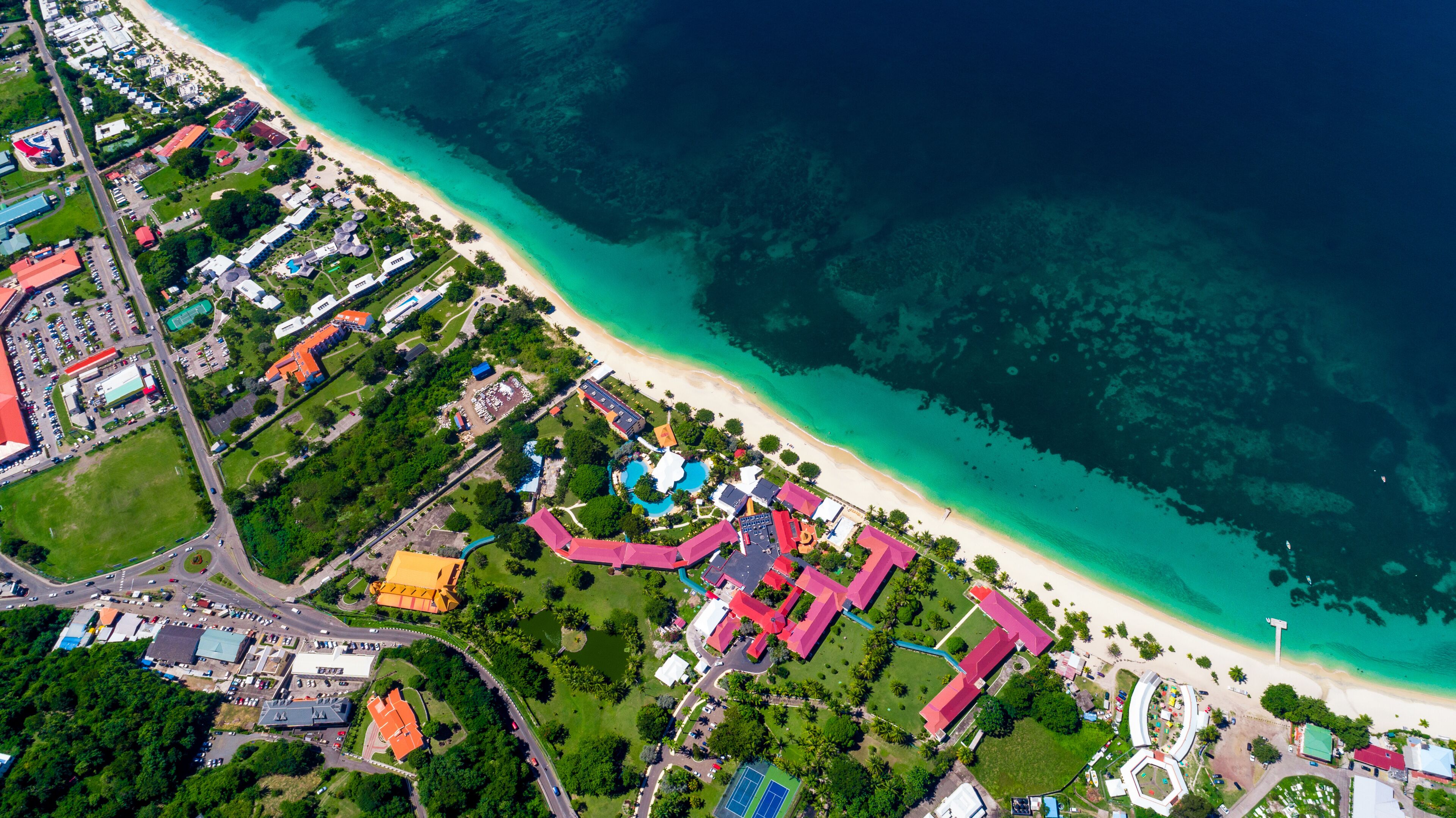 An aerial view of a coastal area with a beach, clear turquoise waters, and a mix of residential and commercial buildings.