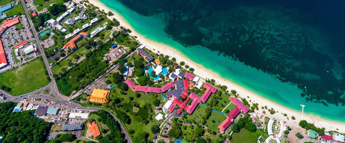 An aerial view of a coastal area with a beach, clear turquoise waters, and a mix of residential and commercial buildings.