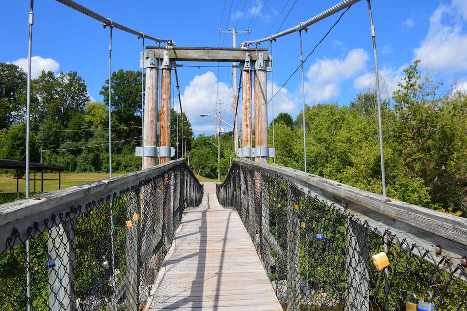A wooden pedestrian bridge with metal railings and a chain-link fence, surrounded by greenery and trees.