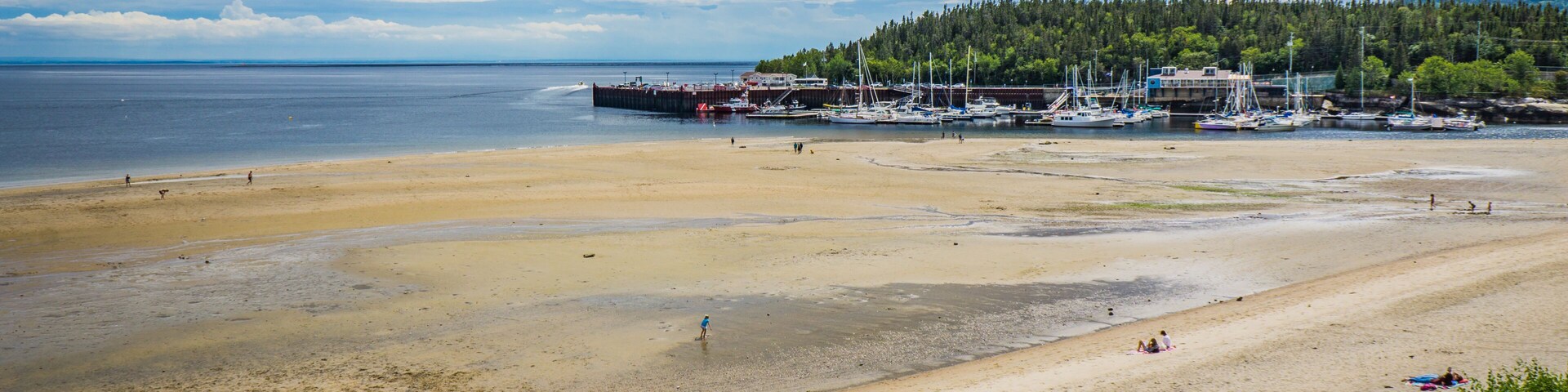 Una playa con gente tomando el sol y nadando, un muelle con barcos, y una colina boscosa al fondo.