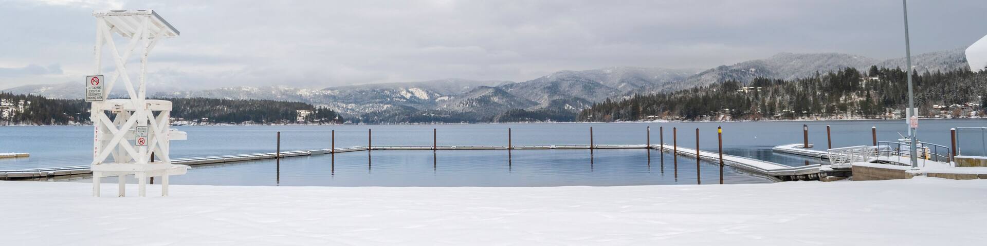 Un paysage hivernal avec une plage enneigée, une tour de surveillance des sauveteurs, un quai et des montagnes en arrière-plan.