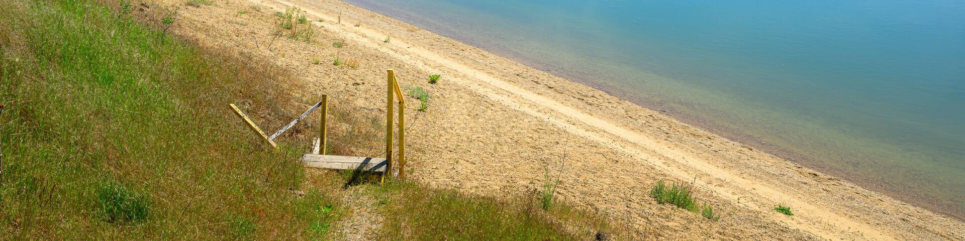 A calm lake with a sandy beach, surrounded by dense forest and a clear sky.