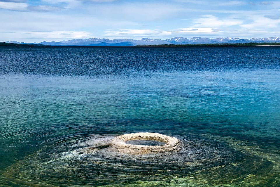 Un lago cristalino con una formación pétrea circular en el centro y montañas al fondo.