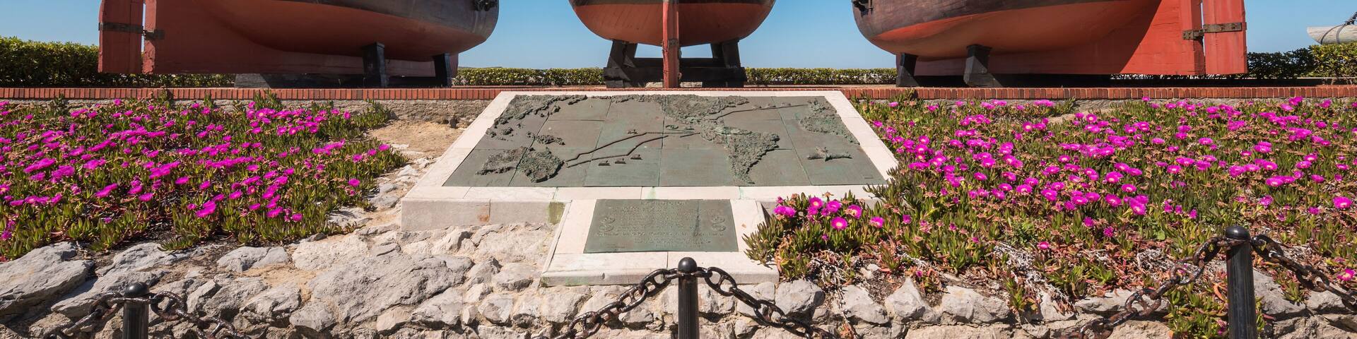 Three large wooden ships with flags, a stone plaque, and pink flowers in the foreground.