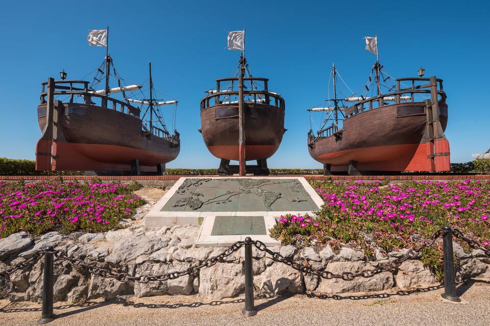 Tres grandes barcos de madera con banderas, una placa de piedra y flores rosadas en primer plano.