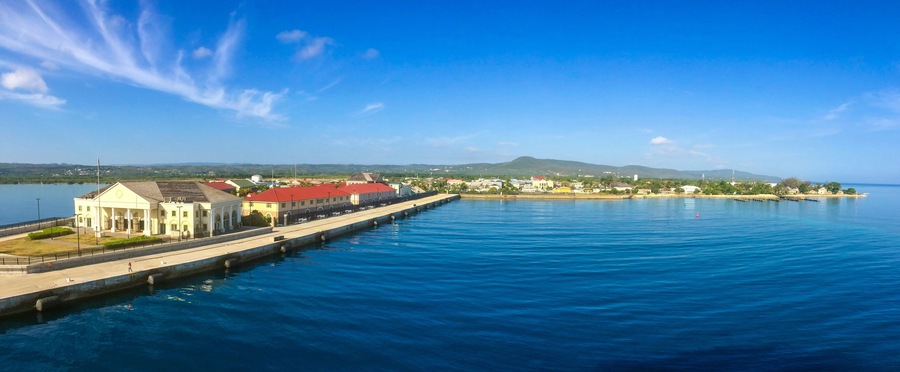 A waterfront promenade with a large white building with a red roof, a pier extending into the water, and a distant mountain range.