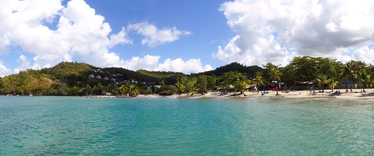 A coastal beach with clear turquoise waters, a sandy shore, and a hill with buildings and palm trees.