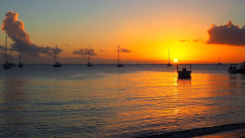 A sunset over a calm sea with several sailboats and a small boat anchored near the shore.