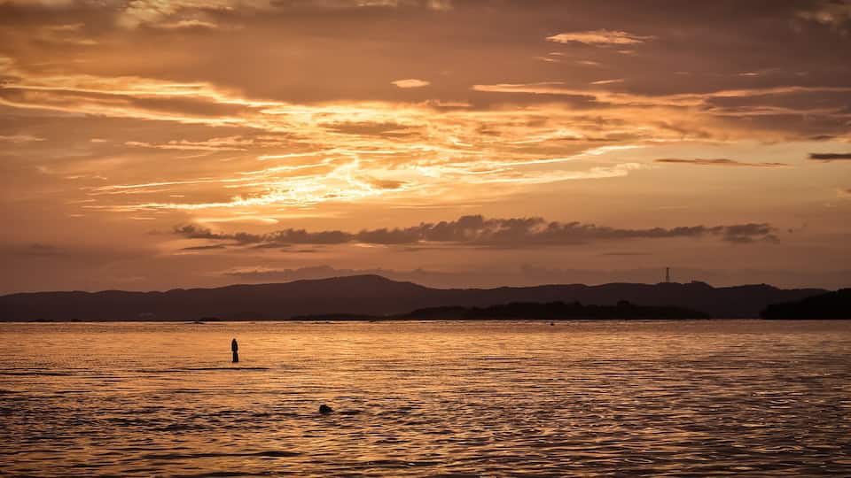 A sunset over a calm body of water with a silhouette of a person standing in the distance and mountains in the background.