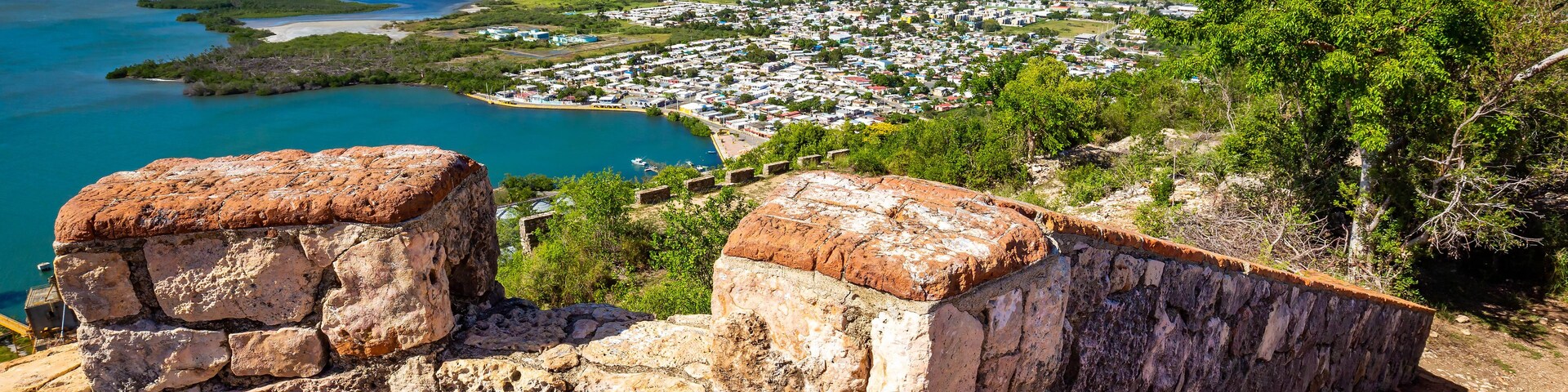 Uma vila costeira com uma muralha de pedra histórica, uma baía com barcos e montanhas ao fundo.