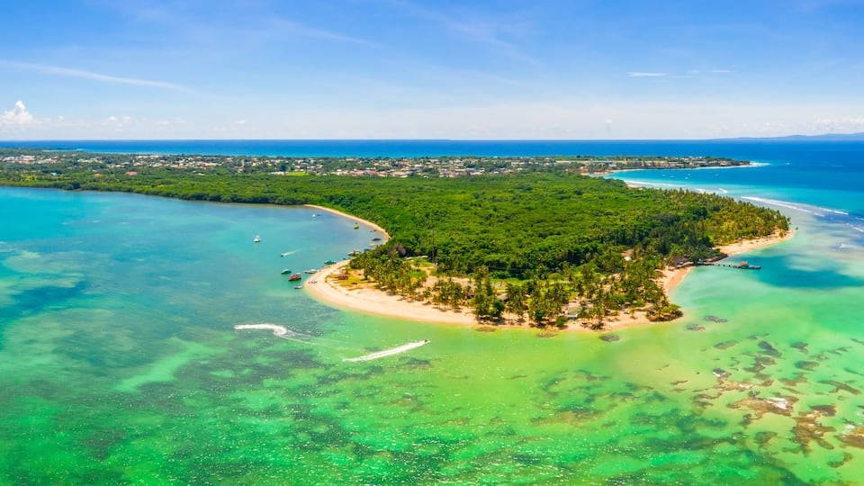 An aerial view of a tropical island with clear turquoise waters, a sandy beach, and dense green vegetation.