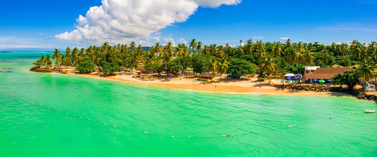 A tropical beach with clear turquoise waters, palm trees, and small huts.
