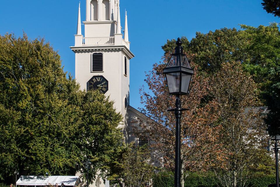 A church with a tall steeple, surrounded by trees and a park with people and a street lamp.