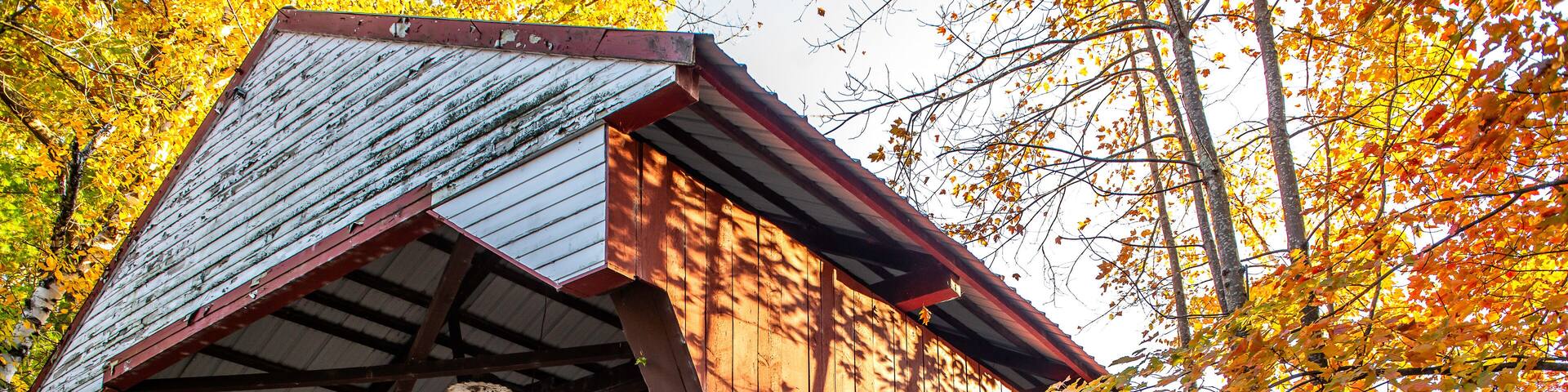 A wooden covered bridge with a metal roof, surrounded by autumn foliage.