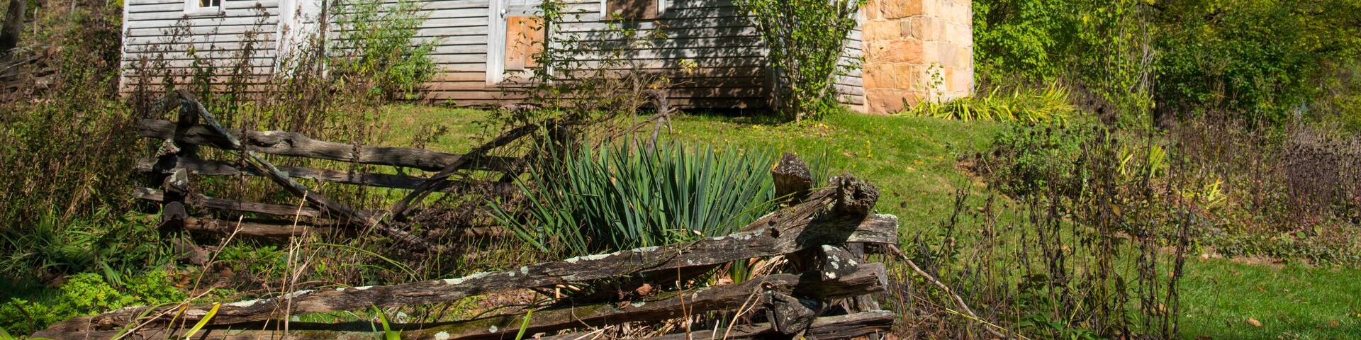 Una casa de madera histórica con chimenea de piedra, rodeada de abundante vegetación y una cerca de madera.
