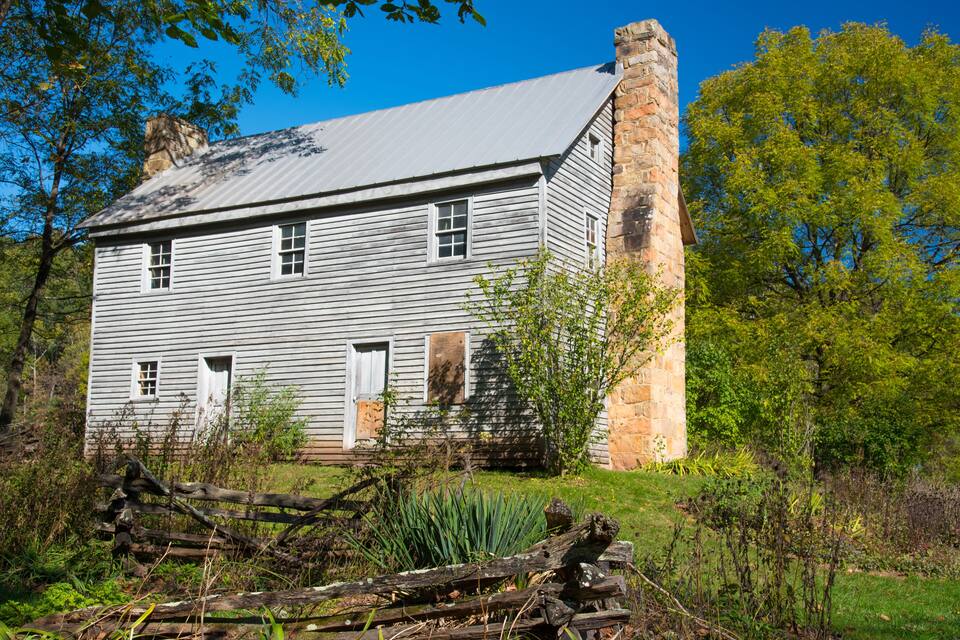 Una casa de madera histórica con chimenea de piedra, rodeada de abundante vegetación y una cerca de madera.