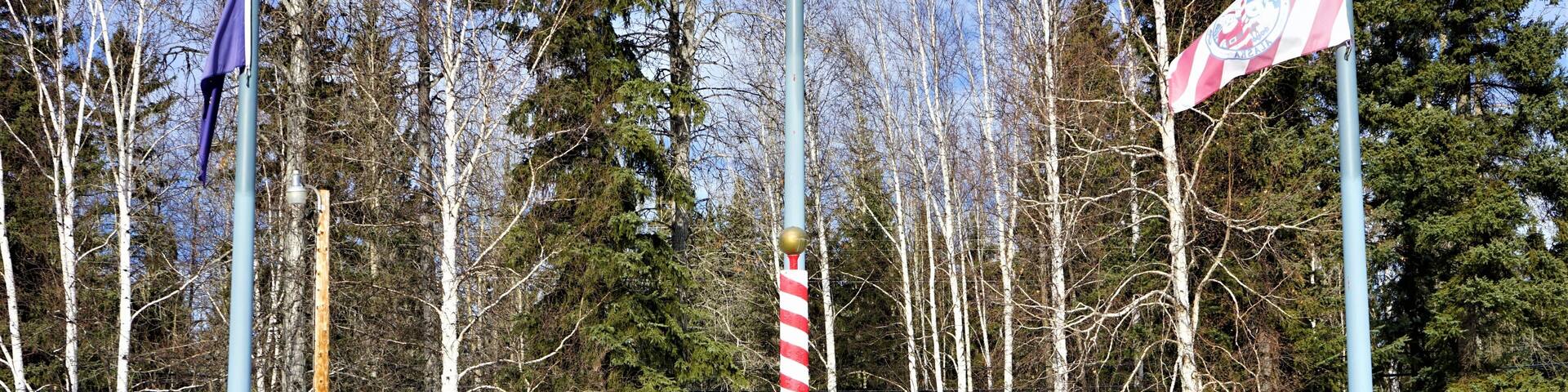 A snowy park with flagpoles, a wooden bench, and a signpost.