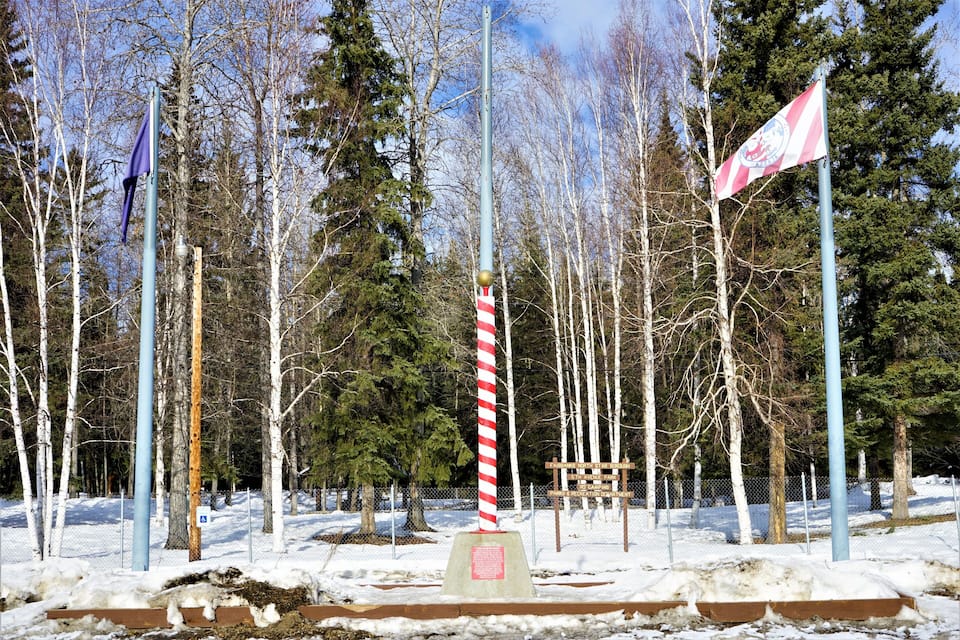A snowy park with flagpoles, a wooden bench, and a signpost.