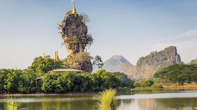 A rock formation with a golden temple on top, surrounded by lush greenery and a calm lake.