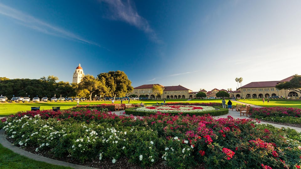 A campus with a large building, a tower, and a well-maintained garden with flowers.